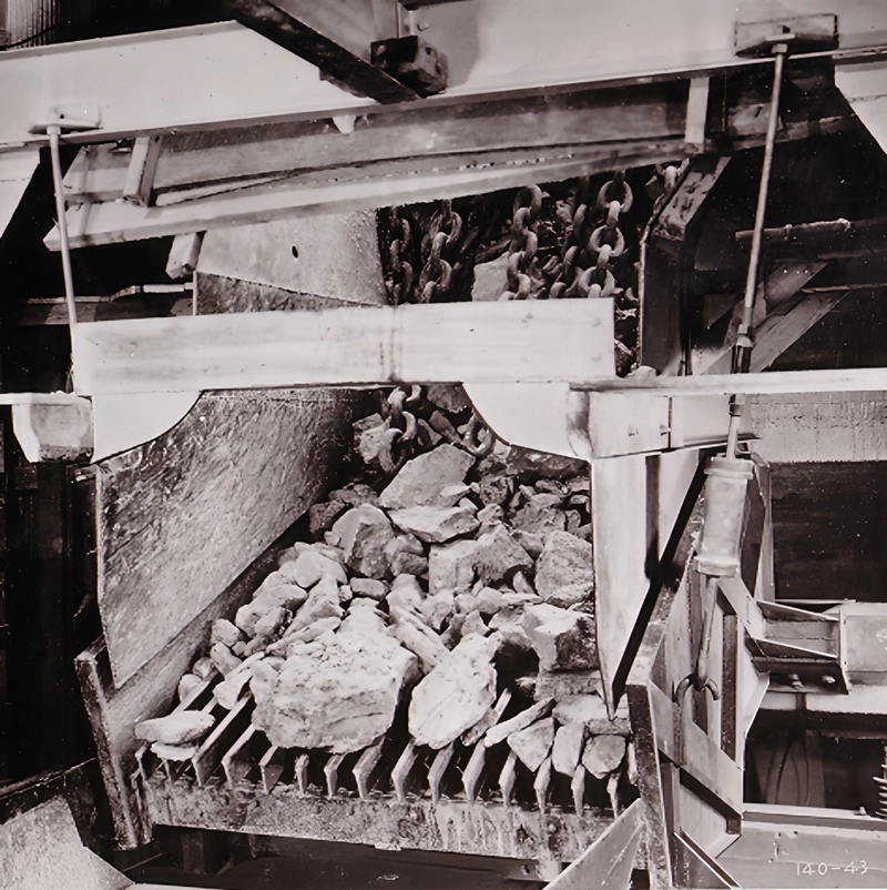 Historic black-and-white photograph of a Jeffrey Rader® electromagnetic vibrating feeder handling large rock material beneath an industrial hopper and chain conveyor system.